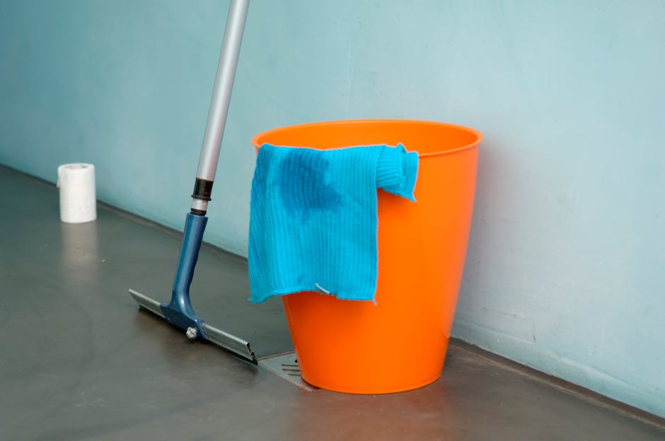 A female cleaner in a kitchen, dressed in a gray t-shirt and red cleaning overalls, holding a white cloth in her right hand and resting her left hand on a blue cleaning bucket. The kitchen has a moder