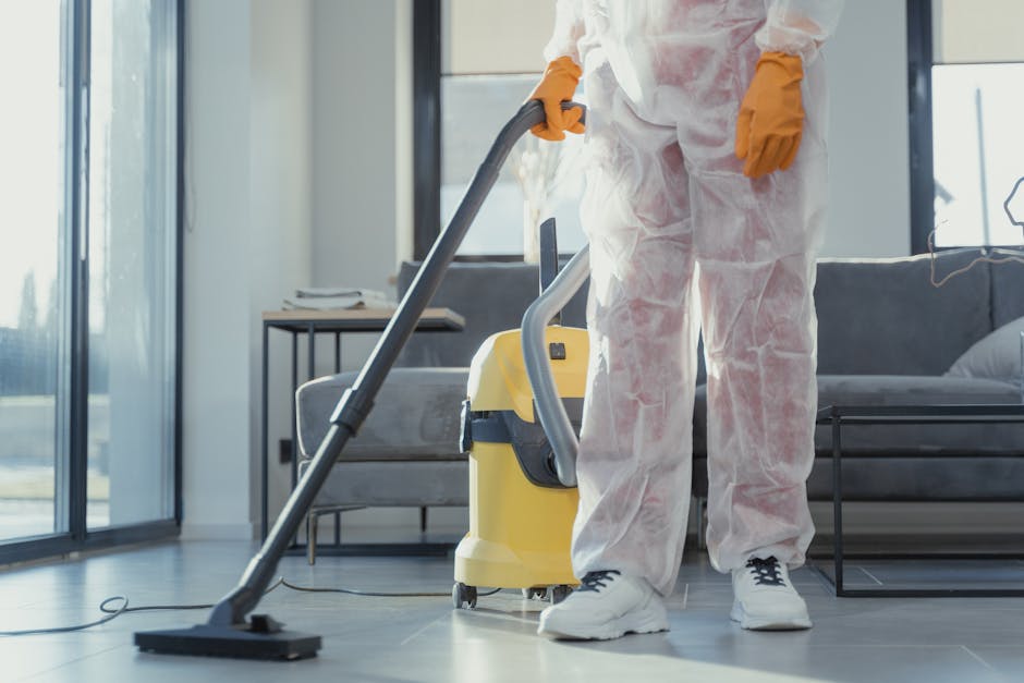 A woman dressed in a dark uniform, likely a cleaning professional, is holding an orange cushion in a well-lit living room. The room features a wooden floor, a partial view of a white wall, and a doorway leading to another area. In the background, there is a bicycle and some household items, indicating a domestic setting. The woman appears to be preparing the cushion for cleaning or repositioning it. The scene emphasizes a clean, tidy space with smooth, polished wooden surfaces and a modern interior design. This image likely relates to comprehensive domestic surface cleaning and maintenance services provided by Kingstonuponthamescarpetcleaners.org.uk, as part of their best local cleaning services near Kingston Bridge.
