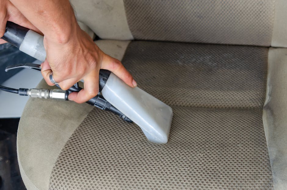 A close-up view of a professional cleaner's hand using a handheld steam or vacuum device to deep clean a beige upholstered car seat with a textured fabric pattern. The cleaning device is pressed onto the fabric, releasing cleaning solutions or steam to remove dirt and stains from the surface. The surrounding area of the seat appears to be slightly damp, indicating an active cleaning process. Visible are the person's hand, part of their arm, and the hose attached to the cleaning machine. The scene takes place indoors, possibly on a vehicle or in a domestic setting, with bright lighting highlighting the cleanliness and the details of the fabric. This process exemplifies surface cleaning and sanitisation, carried out by professional cleaning specialists to maintain hygiene and improve appearance, associated with Kingston Upon Thames Carpet and Upholstery Cleaning Canbury Gardens Kingston KT1, as represented by [COMPANY_NAME].