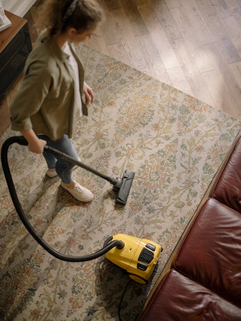 A woman performing surface cleaning with a vacuum cleaner on a patterned area rug in a living room. The rug features intricate floral designs in muted tones and is placed on a wooden floor. She is wearing casual clothing, including a green jacket and white sneakers. The room is well-lit, showcasing a leather sofa adjacent to the rug. This scene illustrates deep cleaning and sanitisation practices carried out by Kingstonuponthamescarpetcleaners.org.uk for maintaining hygienic home environments.